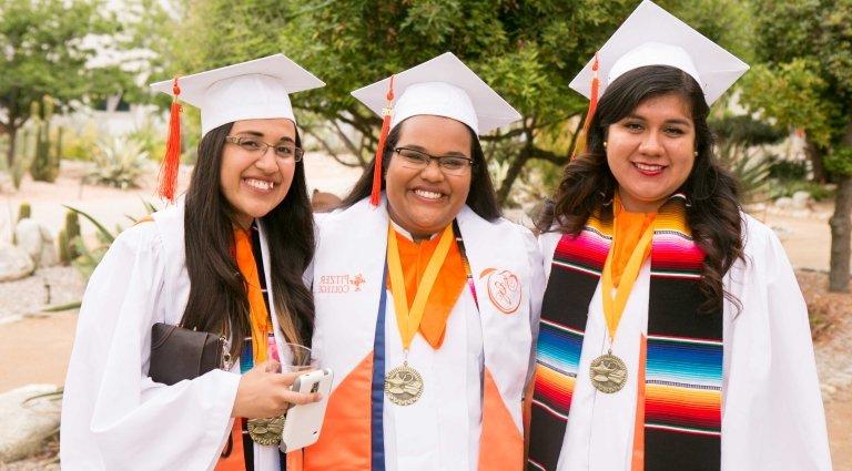 Three female graduates in Scott Courtyard, two with Chicano/Latino stole and one with CAPAS stole. Karen Kandamby is in the center; others are unidentified.